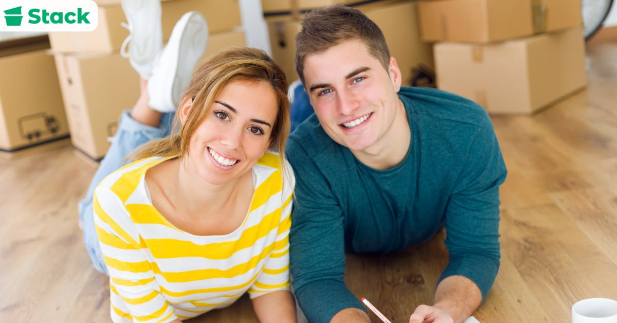 Smiling couple lying on the floor of their new home surrounded by stacked cardboard moving boxes, writing on a notepad