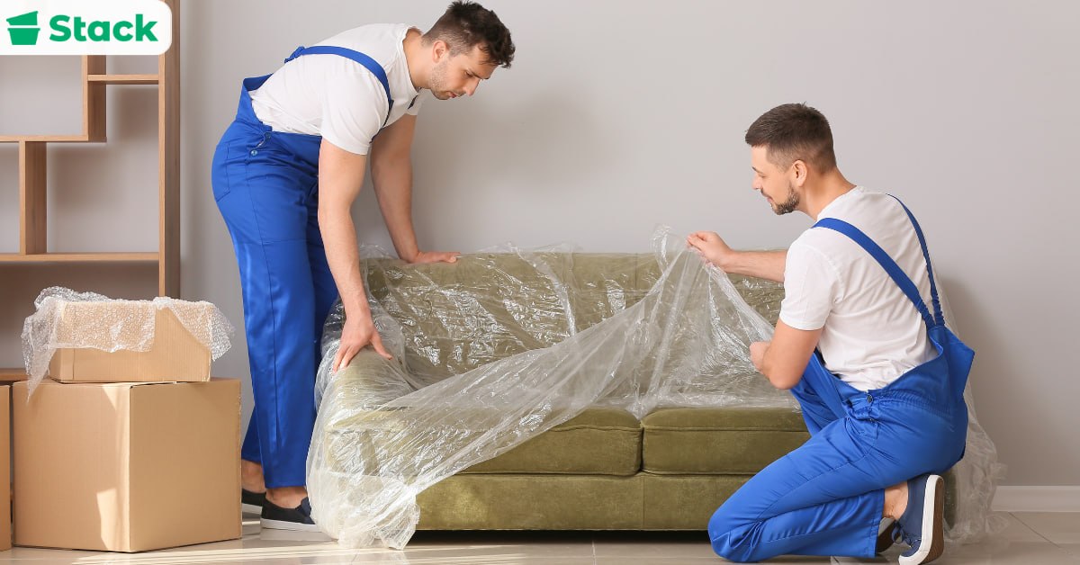Two movers in blue uniforms wrapping a green sofa in plastic stretch film, with bubble-wrapped cardboard boxes stacked beside them